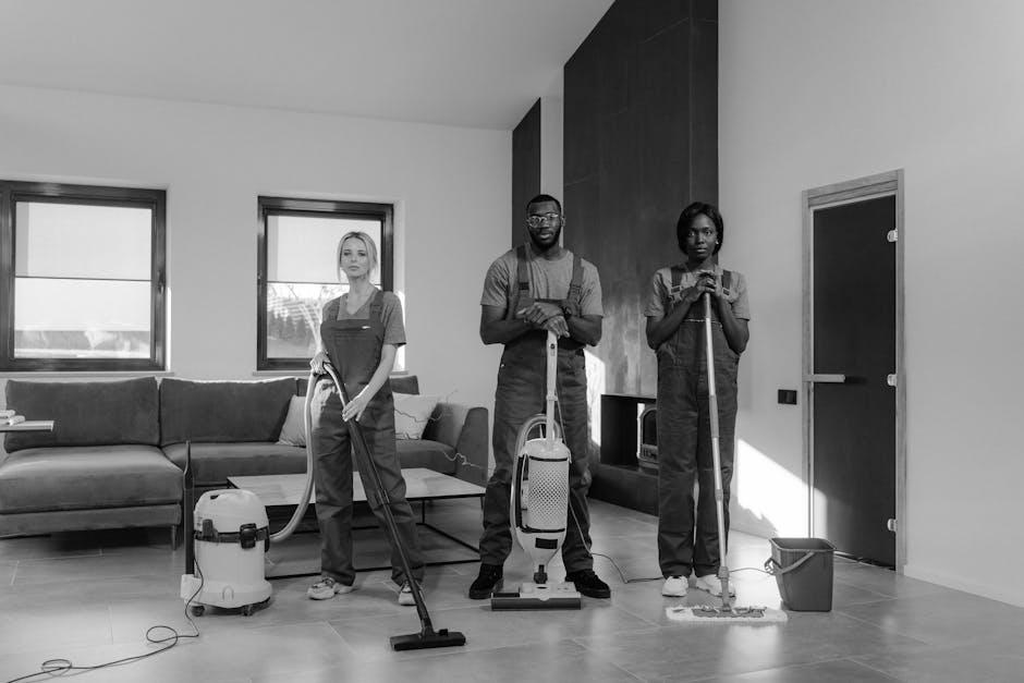 Three professional cleaners standing in a modern living room with light-colored wooden flooring, equipped with vacuum cleaners and mops, preparing for deep surface cleaning and sanitisation. The room