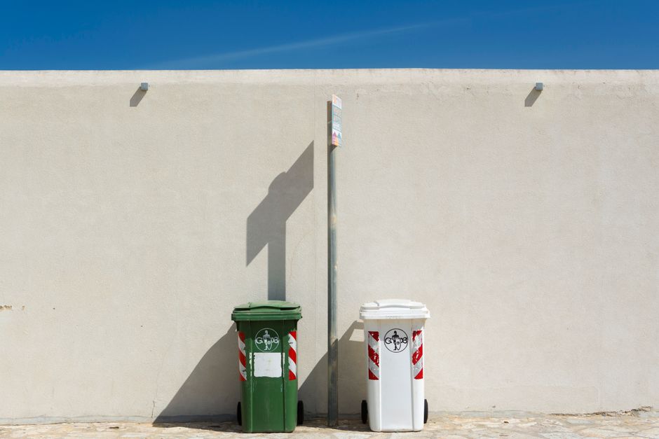 Two waste disposal bins, one green and one white, positioned outdoors against a plain beige wall, with a metal pole between them. The green bin, situated on the left, has a lid and a label area that a