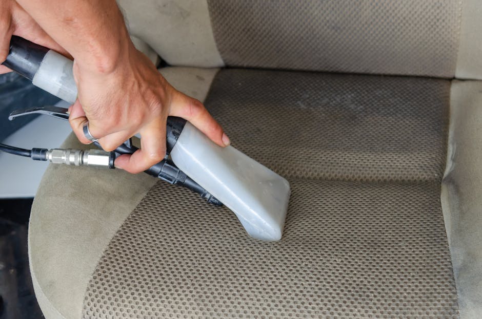 A person's hand holding a handheld upholstery cleaning machine, using it to deep clean a beige fabric car seat with a textured pattern. The device has a gray body with a black hose connected to a tran
