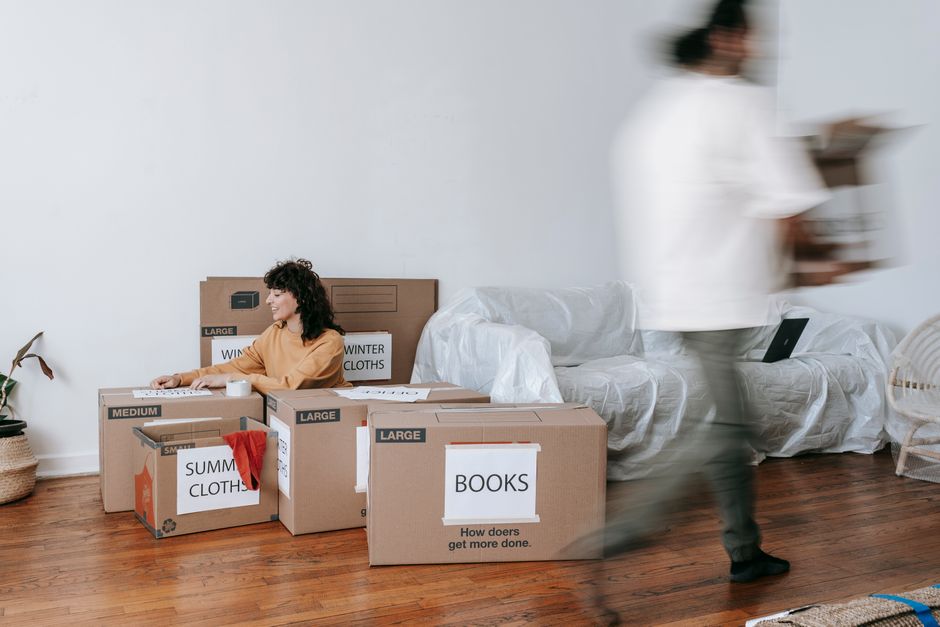 A woman with curly dark hair, dressed in a beige top, is sitting on a white sofa that is covered with a protective sheet, surrounded by large cardboard boxes labeled 'SUMMER CLOTHS,' 'BOOKS,' and 'WIN