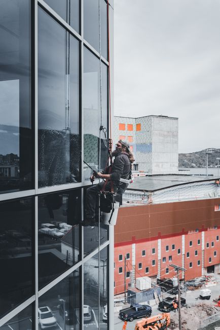 Two professional window cleaners wearing orange safety harnesses and helmets are suspended on a tall, modern glass-fronted office building. They are cleaning the large, reflective glass panels from th